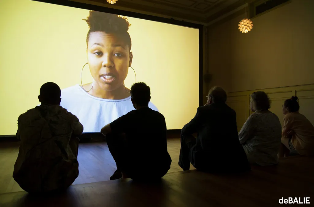 Publiek kijkt naar een grote videoprojectie met een persoon met een donkere huidskleur die naar de camera kijkt voor een felgele achtergrond.
