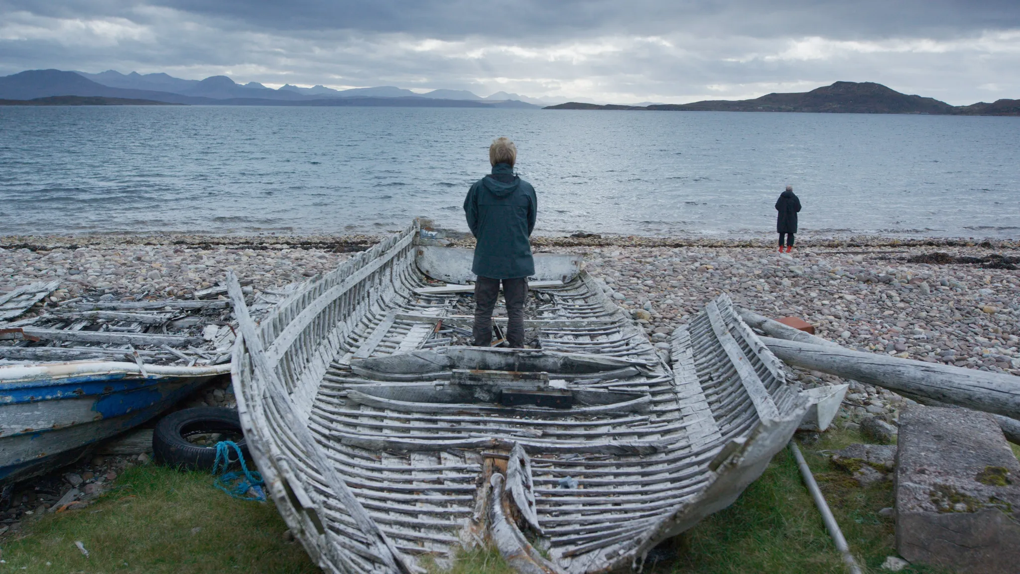 Een persoon staat op een wrak houten schip op een kiezelstrand, starend naar het water