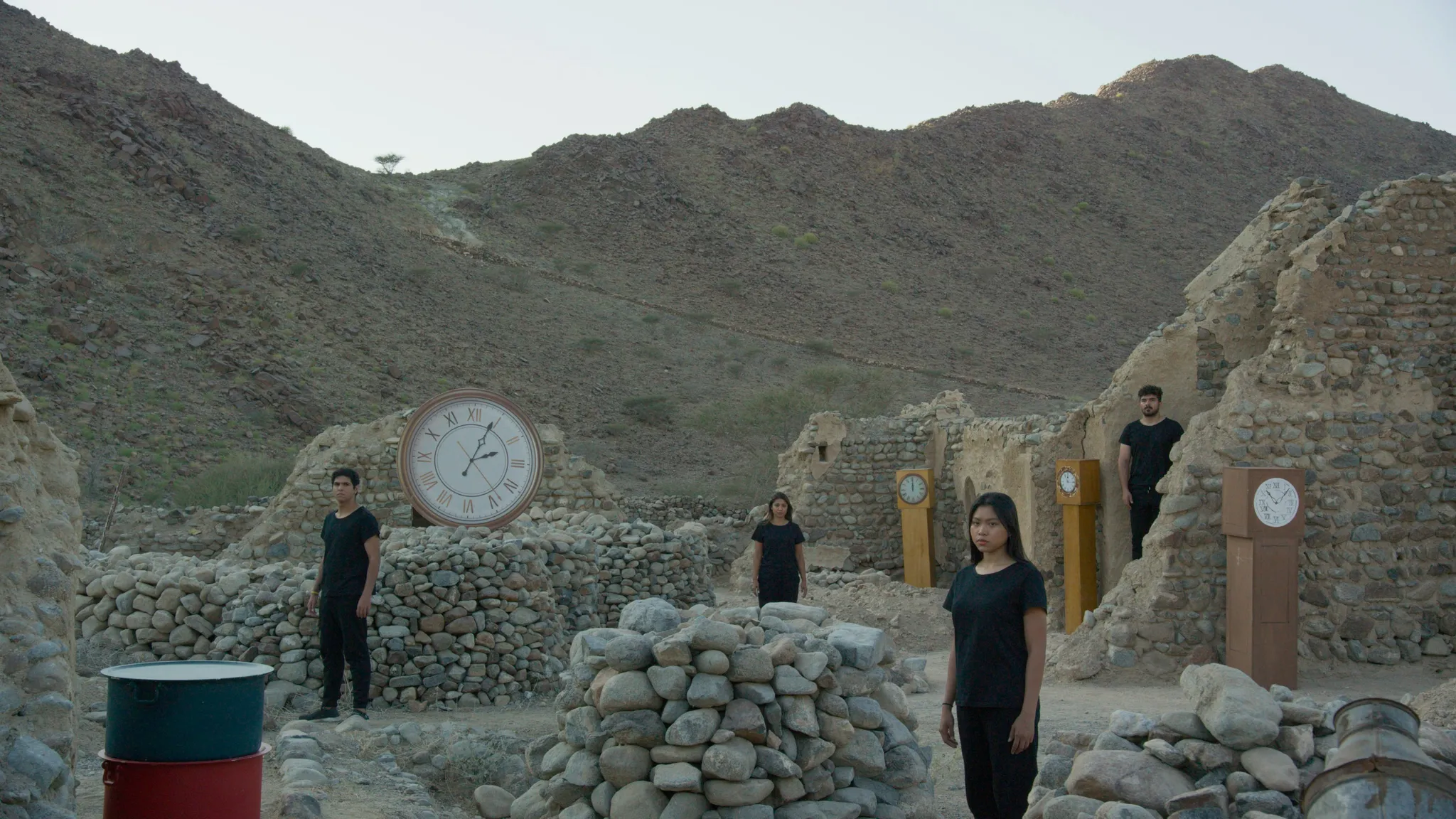 4 in het zwart geklede personen staren naar de camera, staande tussen ruïnes en grote klokken, in een verlaten rotslandschap