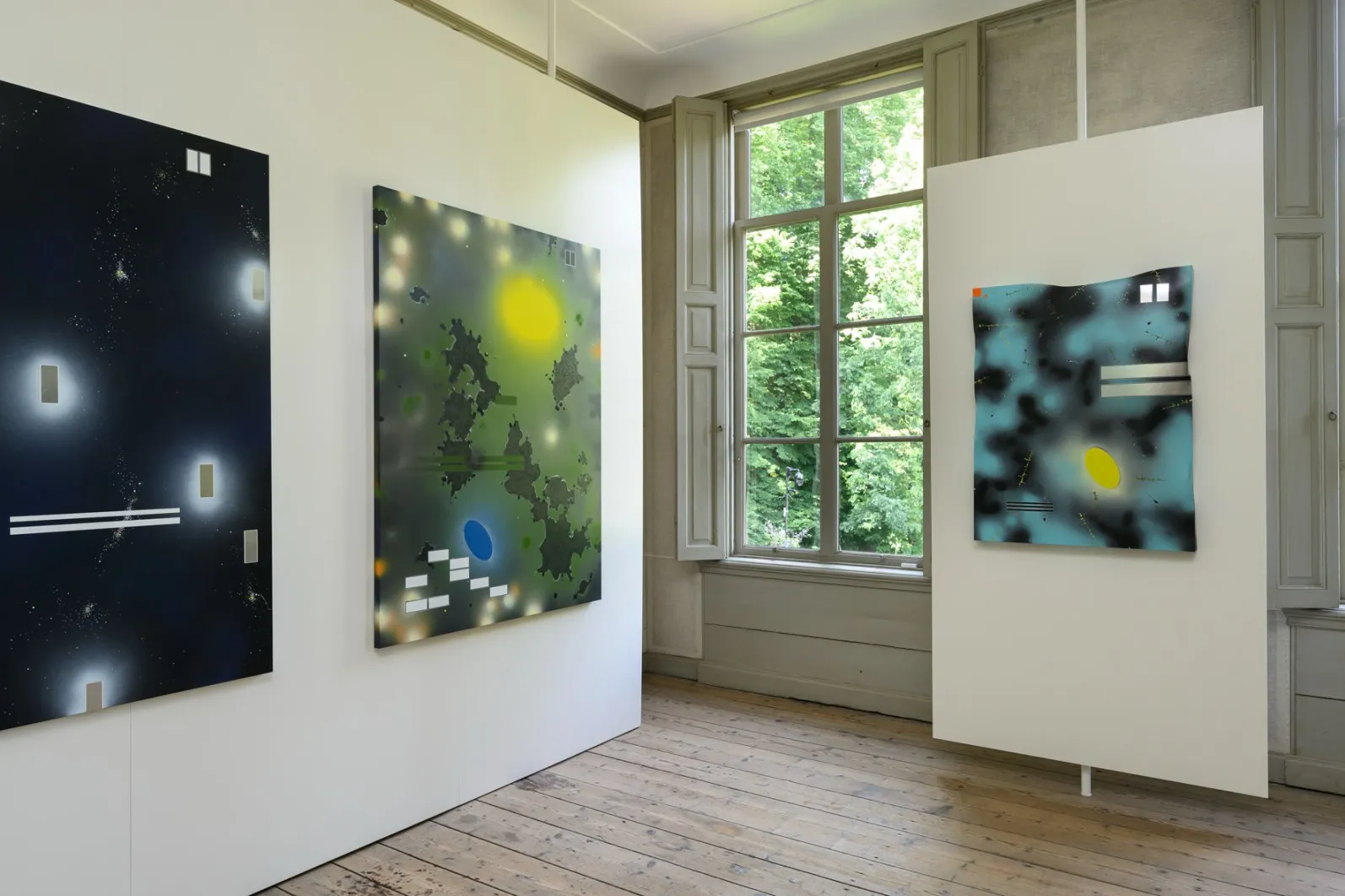Drie abstracte kunstwerken bij een raam, badend in natuurlijk licht, waar bomen van buitenaf zichtbaar zijn, in de expositieruimte.