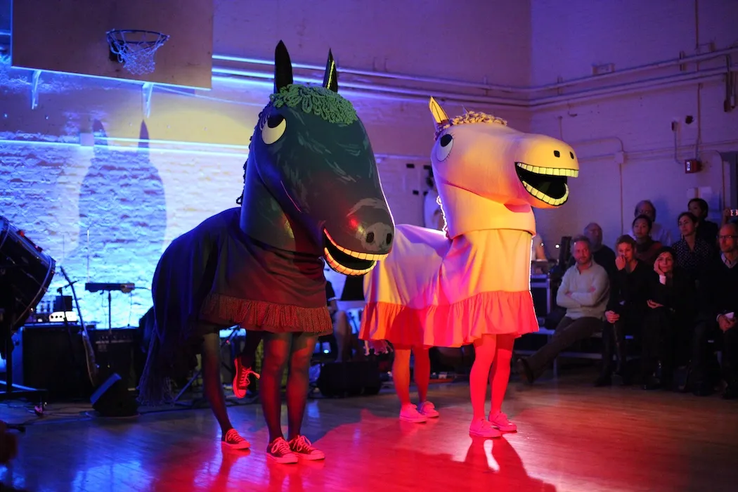 Two costumes of smiling cartooney horses with performers underneath, performing in front of a public on a basketball field