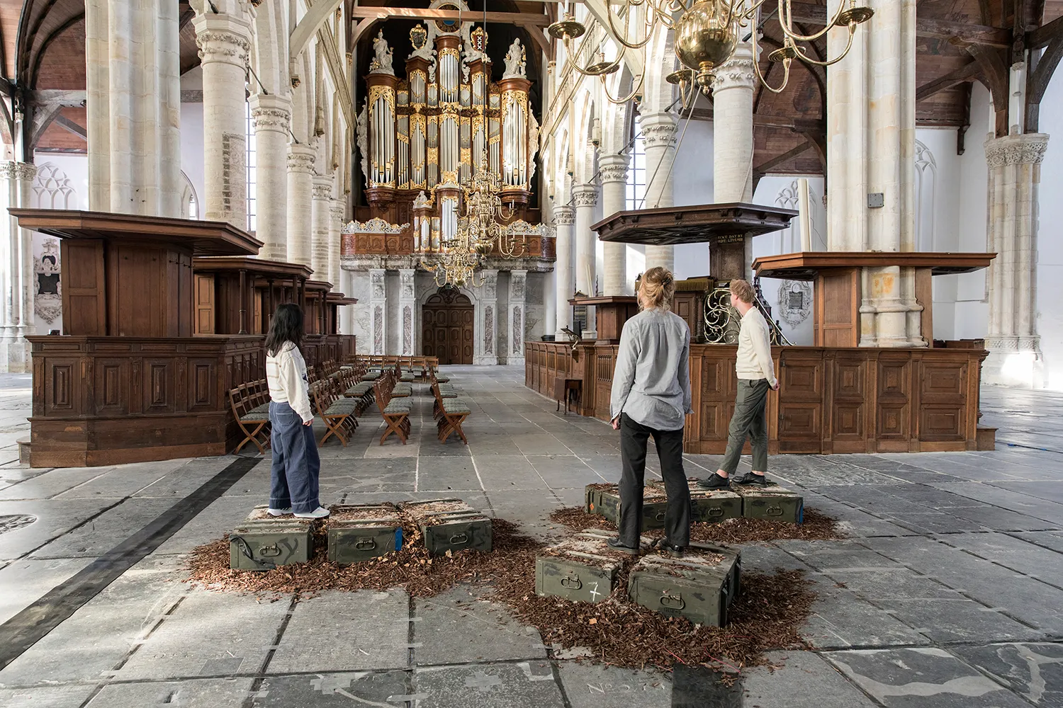 Drie personen staand op groene houten kisten geplaatst op houtsnippers, starend naar het kerkorgel in de Oude Kerk
