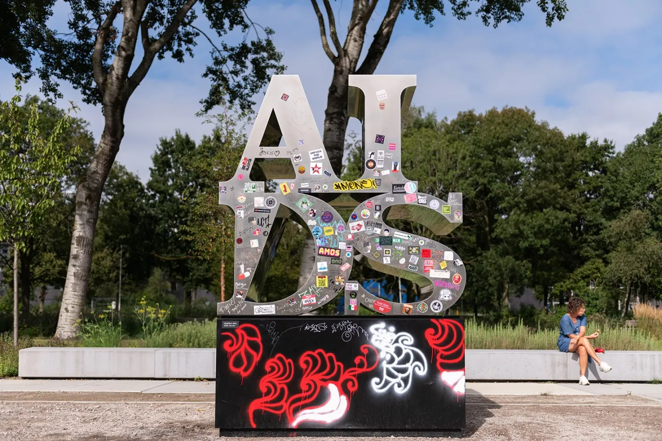 A and I letters on top of D and S silver metal letters (reads AIDS) adorned with stickers and other graffitis placed on a black pedestal in a park