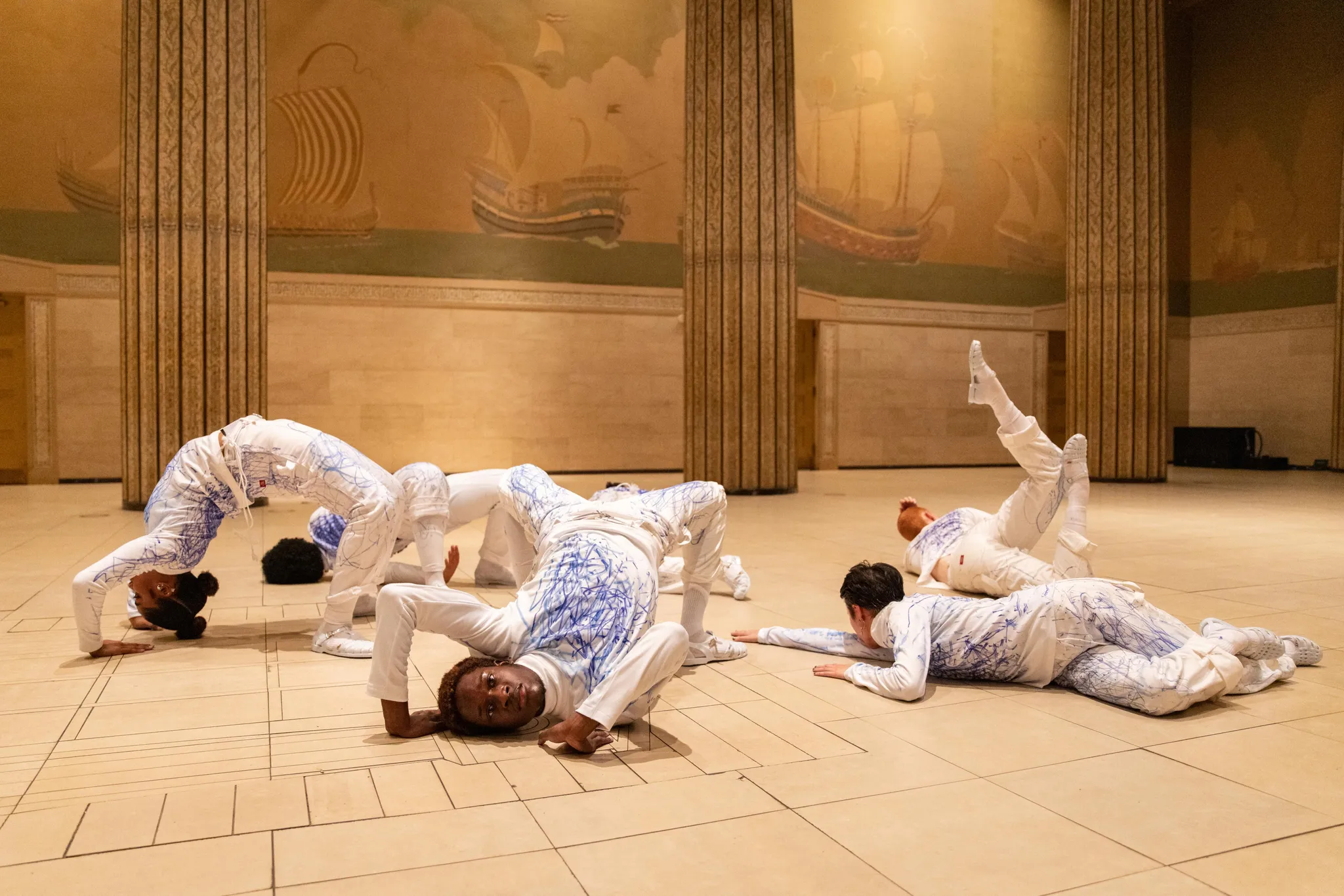 Zes performers in verschillende posities op de vloer, midden in een grote balzaal met pilaren en muurschilderingen van schepen