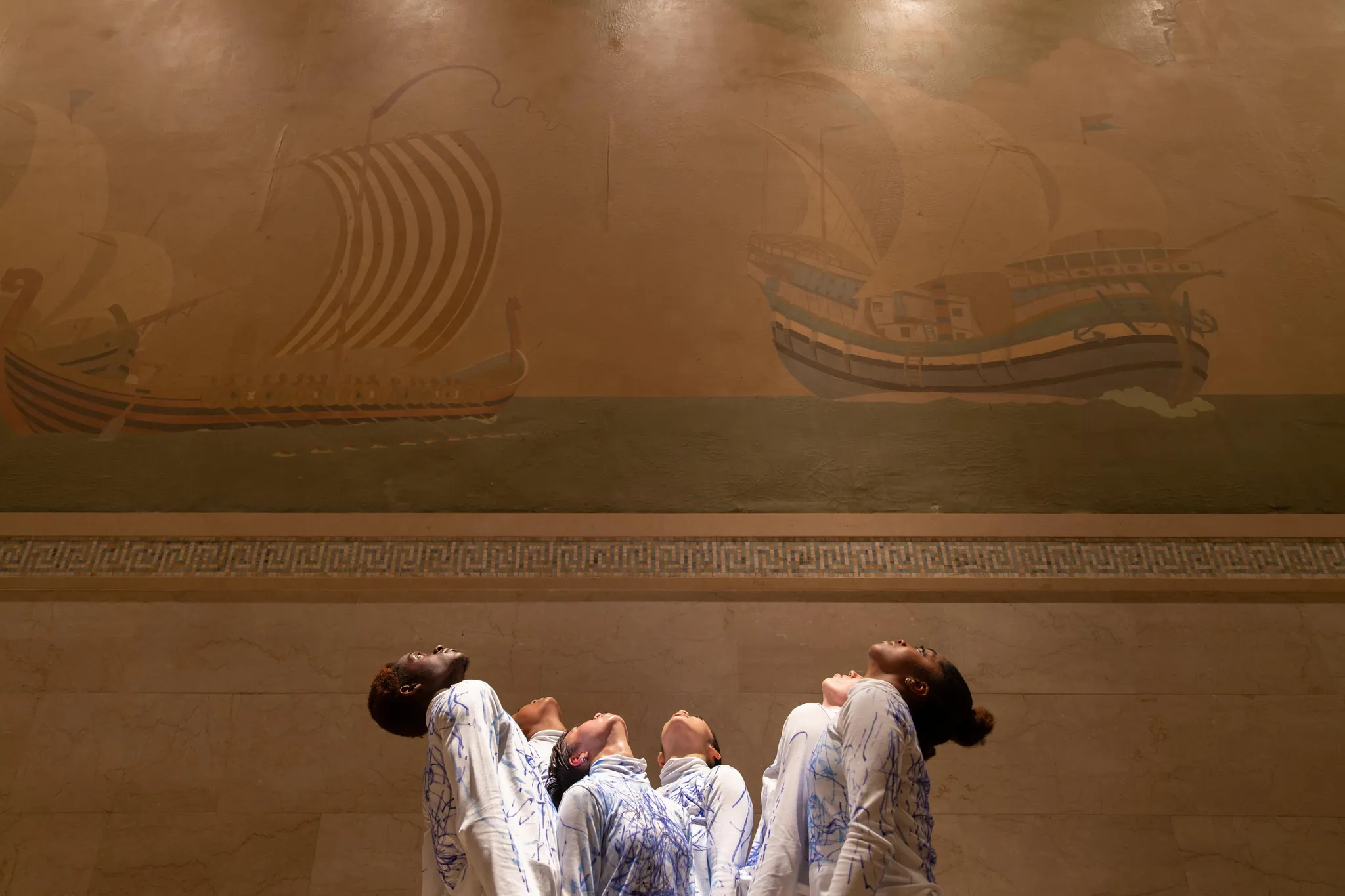 Zes performers van verschillende etnische achtergronden kijken omhoog, voor een muurschildering van twee oude schepen