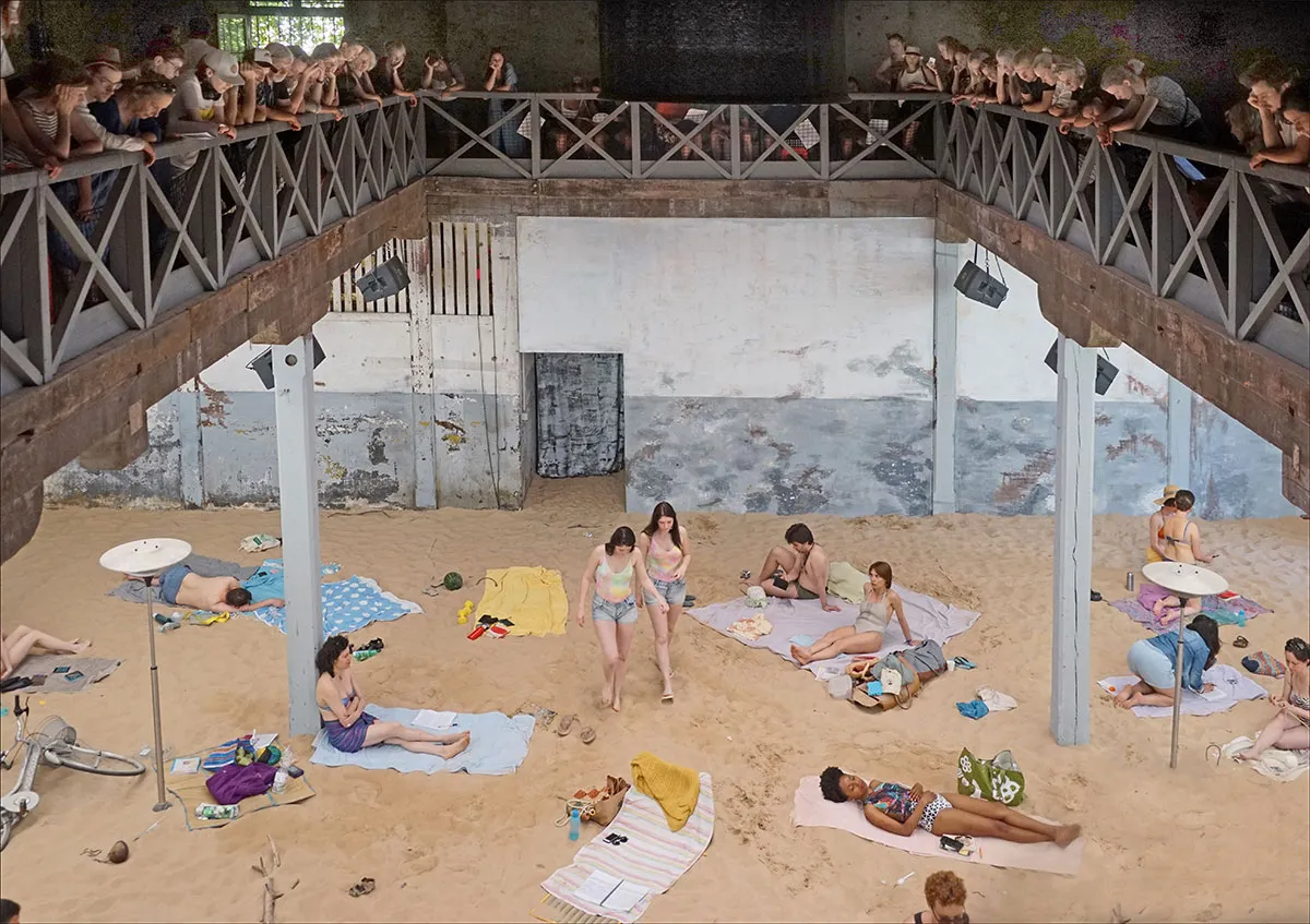 Het publiek kijkt neer vanaf een balkon rondom boven de performers die met kleurrijke handdoeken en strandaccessoires op het kunstmatige strand liggen.