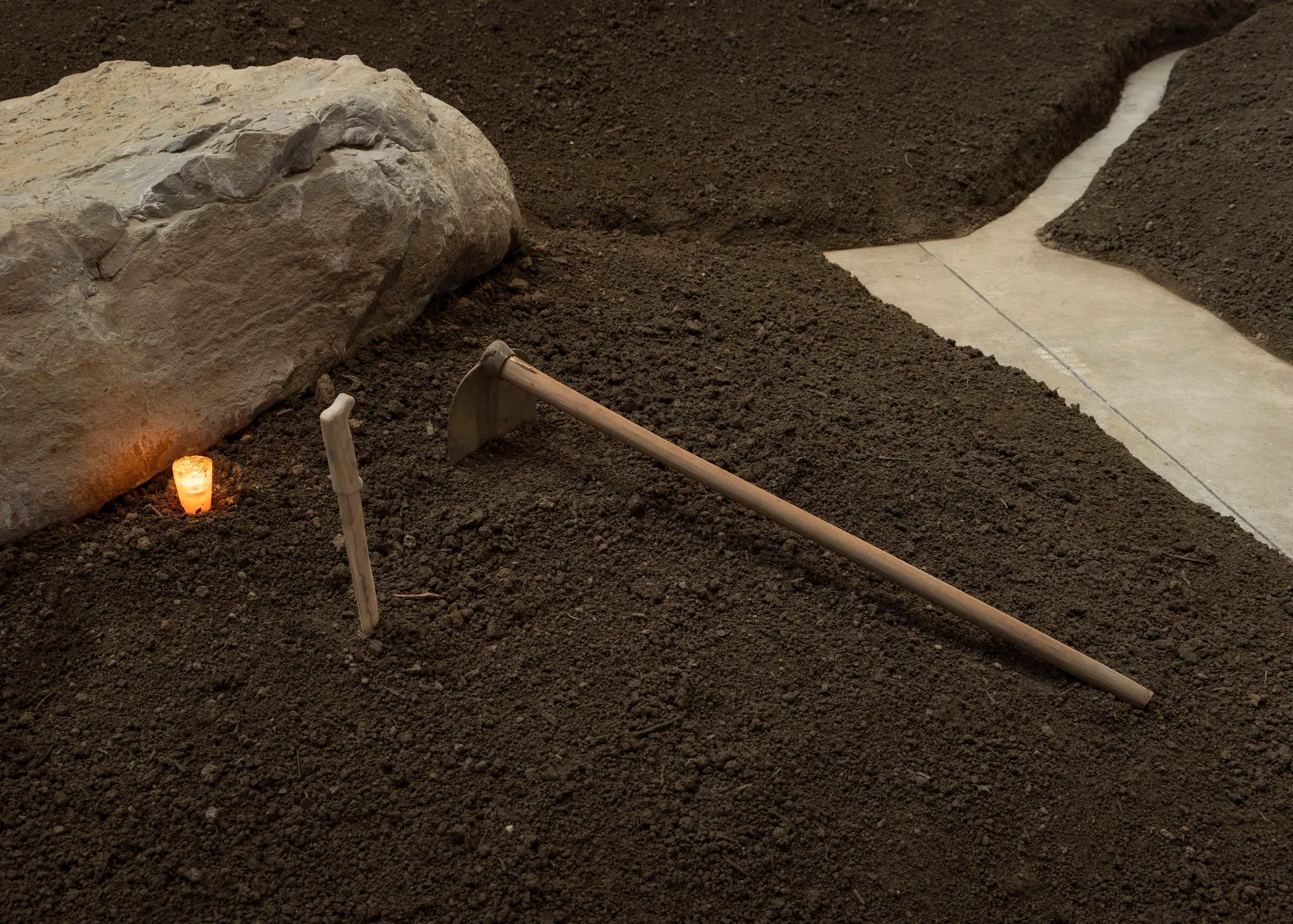 Close up van de installatie: een steen, een kaars, een houten mes en een schoffel op de bruine aarde