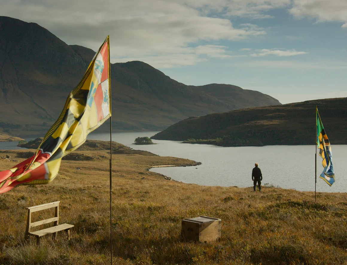 A mountainous landscape with a lake (or river), two undefined flags planted in the ground and a person staring at the water