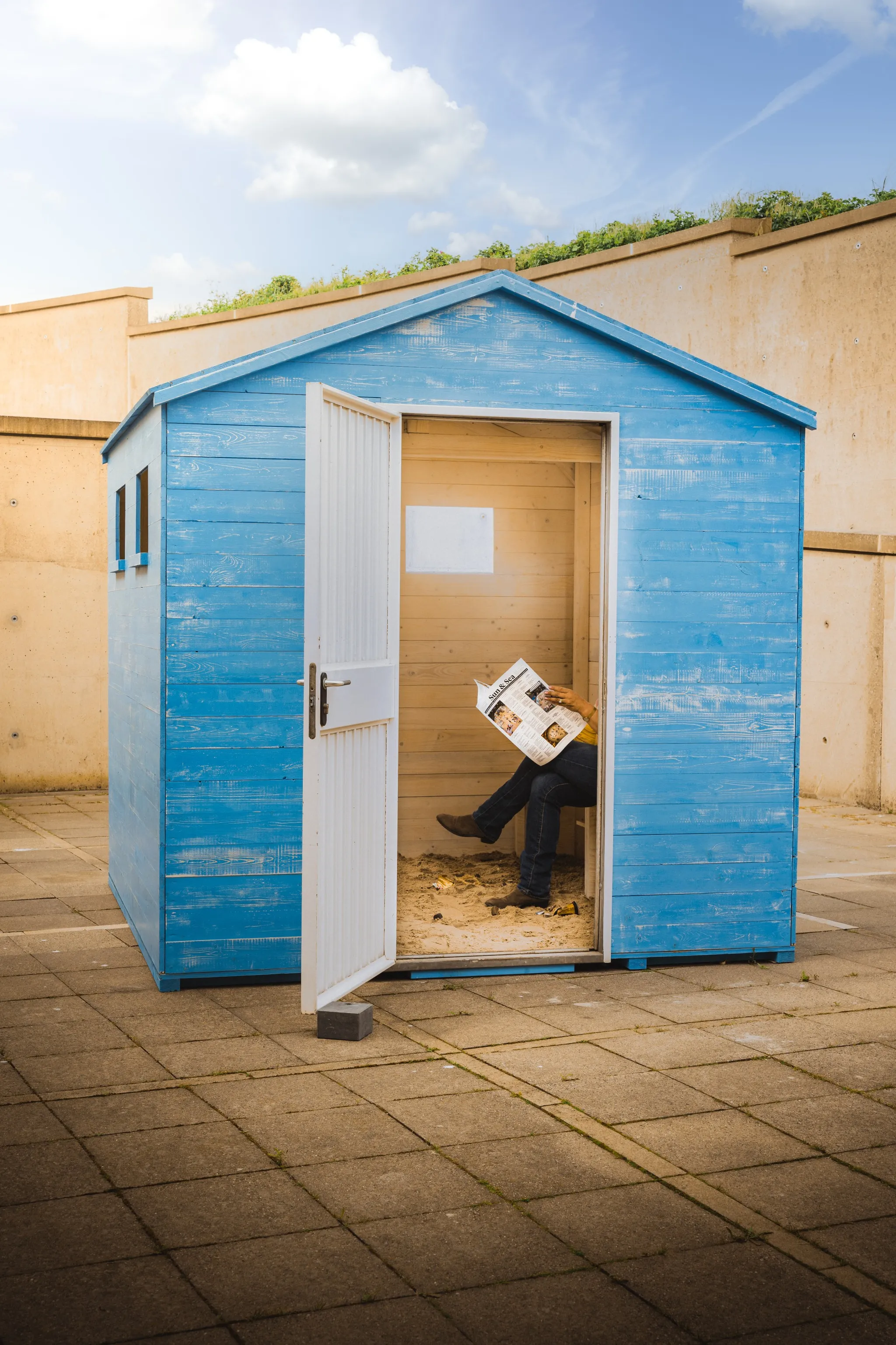 A bright blue wooden beach cabin sits on a concrete floor. Inside, a visitor reads a newspaper.